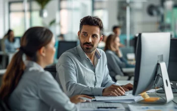 A lifestyle stock photography of a professional helpdesk technician, seated at a modern office desk, engaging with a colleague while working on a computer; the scene is set in a contemporary, open-plan office space with other employees visible in the background; the composition includes sleek office furniture, computer monitors, and paperwork scattered neatly; the lighting is bright and natural, streaming in from large windows; the framing captures a mid-shot, focusing on the interaction between the technician and the colleague; the mood is collaborative and productive, emphasizing teamwork and efficiency; the camera angle is at eye level, providing a clear view of the technician's face and workspace. --ar 3:2 --no text, fonts, letters, watermark, words, typography, slogans, signature, camera --v 6.0 --s 250